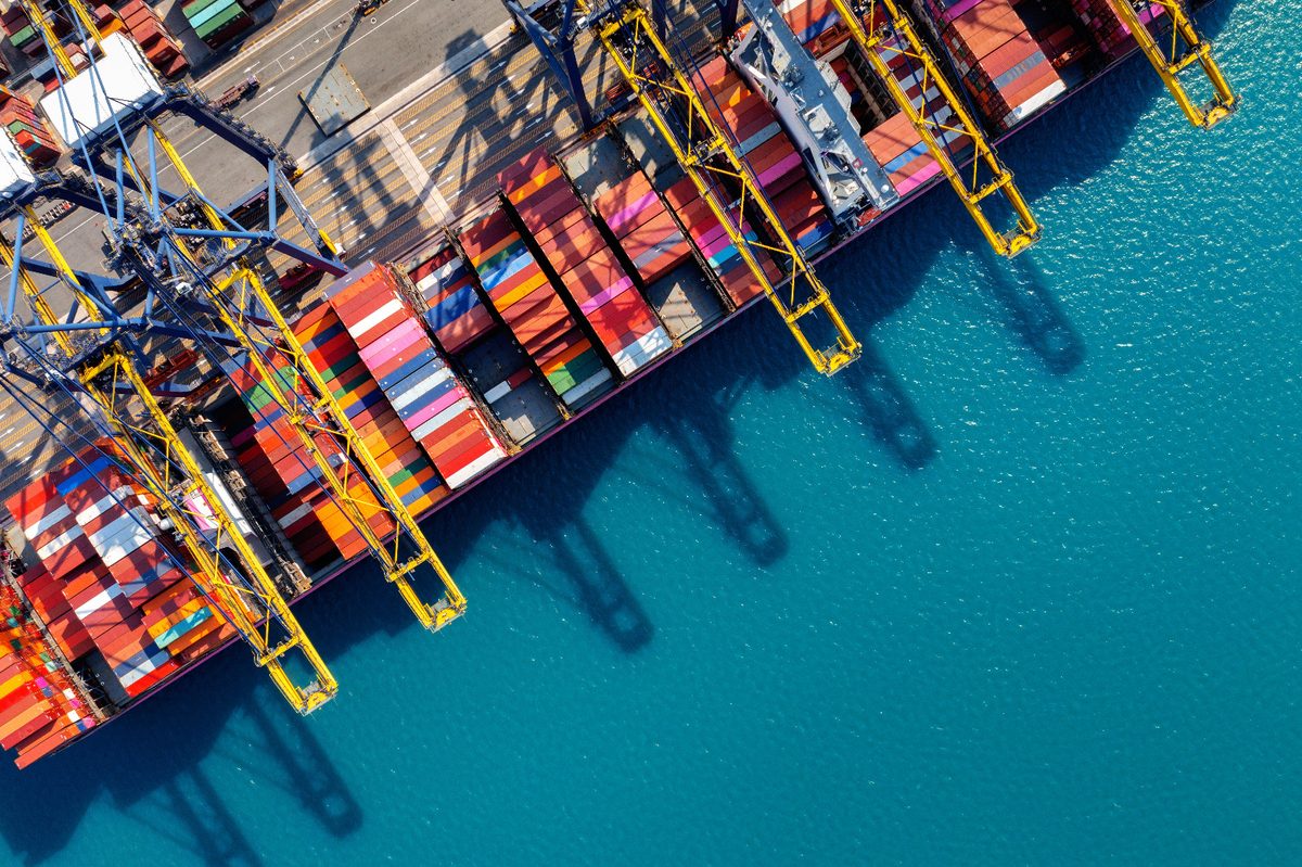 Aerial view of container ships being loaded at a port, with cranes casting long shadows across the water